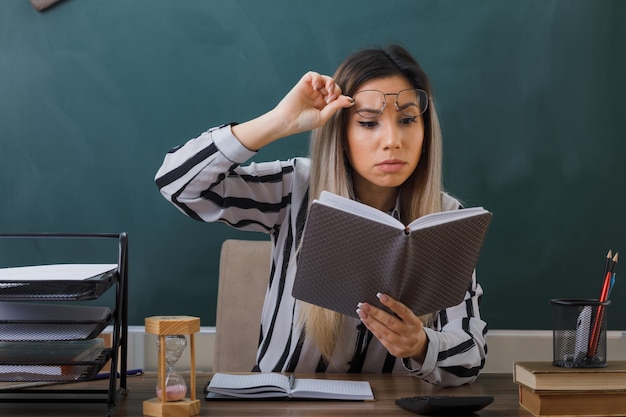 young woman teacher wearing glasses sitting at school desk in front of blackboard in classroom checking homework of students looking surprised and confused