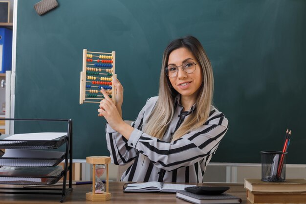 young woman teacher wearing glasses sitting at school desk in front of blackboard in classroom explaining lesson holding abacus smiling confident