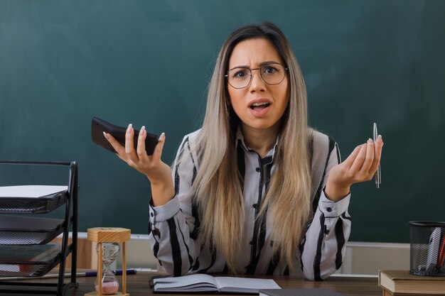 young woman teacher wearing glasses sitting at school desk in front of blackboard in classroom explaining lesson holding calculator looking at camera confused having no answer