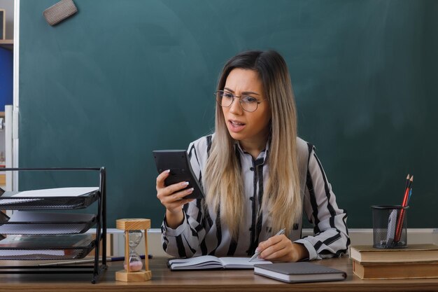 young woman teacher wearing glasses sitting at school desk in front of blackboard in classroom explaining lesson holding calculator looking at it confused