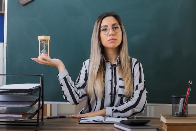 young woman teacher wearing glasses sitting at school desk in front of blackboard in classroom explaining lesson holding hourglass looking confident with serious face