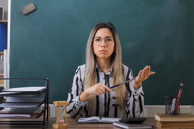 真面目な顔でカメラを見てポインターを保持しているレッスンを説明する教室の黒板の前の学校の机に座って眼鏡をかけている若い女教師