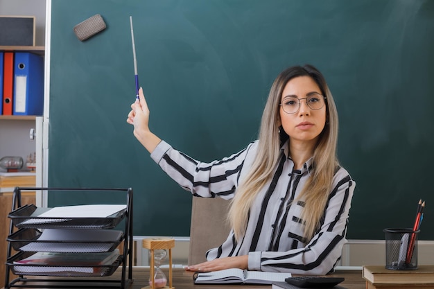 молодая женщина-учитель в очках сидит за школьной партой перед доской в классе, объясняя урок, указывая указкой на доску, выглядит уверенно