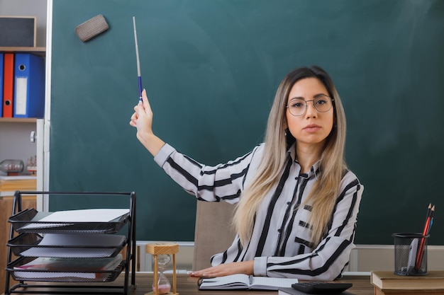 young woman teacher wearing glasses sitting at school desk in front of blackboard in classroom explaining lesson pointing with pointer at blackboard looking confident