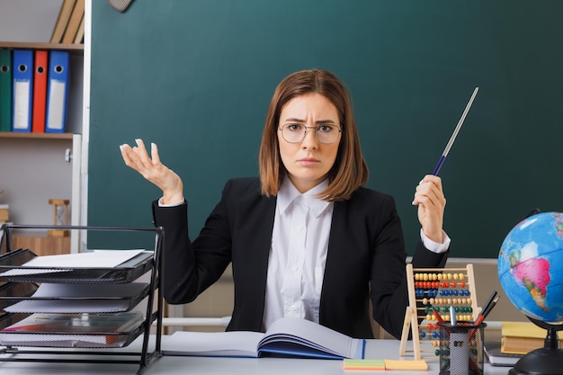 young woman teacher wearing glasses sitting at school desk in front of blackboard in classroom and globe checking class register holding pointer looking displeased raising arm in displeasure