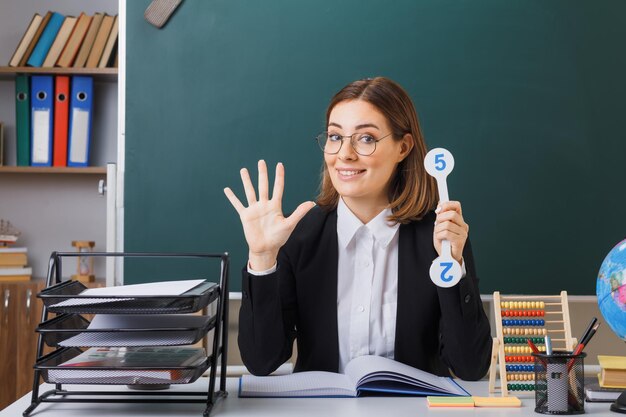 開いた手のひらで5番目を示す幸せで前向きなレッスンを説明するナンバープレートを保持している教室の黒板の前にある学校の机に座っている眼鏡をかけている若い女性教師