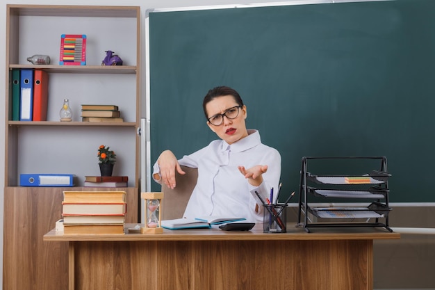 眼鏡をかけた若い女教師が教室の黒板の前の学校の机に座ってカメラを見て不機嫌に腕を上げて不機嫌に