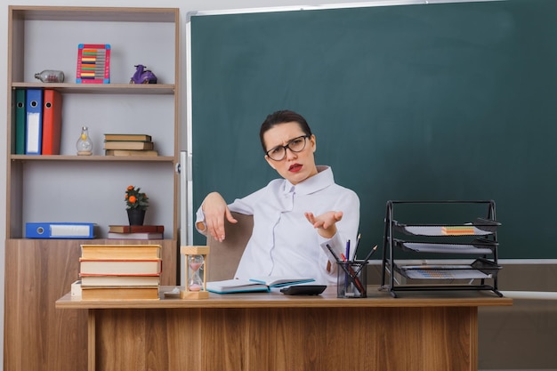 Young woman teacher wearing glasses sitting at school desk in front of blackboard in classroom looking at camera being displeased raising arm in displeasure