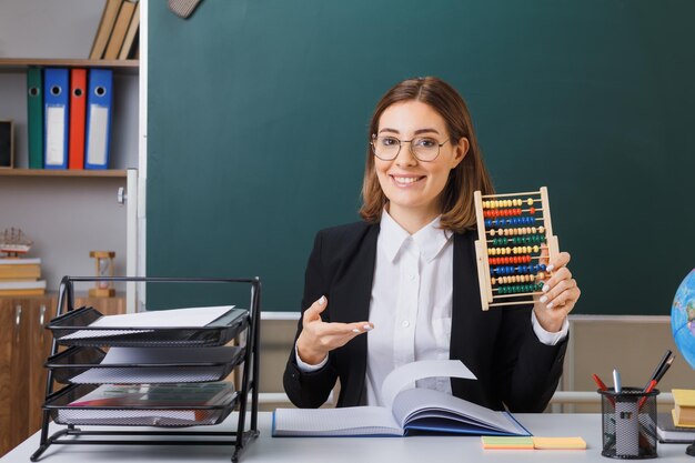 Молодая женщина-учительница в очках сидит за школьной партой перед доской в классе, используя счеты, объясняя урок счастливой и позитивной улыбкой