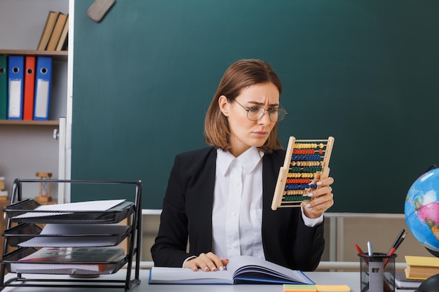 Молодая женщина-учительница в очках сидит за школьной партой перед доской в классе, используя счеты, объясняя урок, хмуро глядя