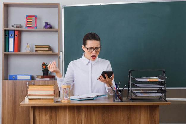 Молодая женщина-учитель в очках сидит за школьной партой перед доской в классе с помощью калькулятора и выглядит смущенной и удивленной