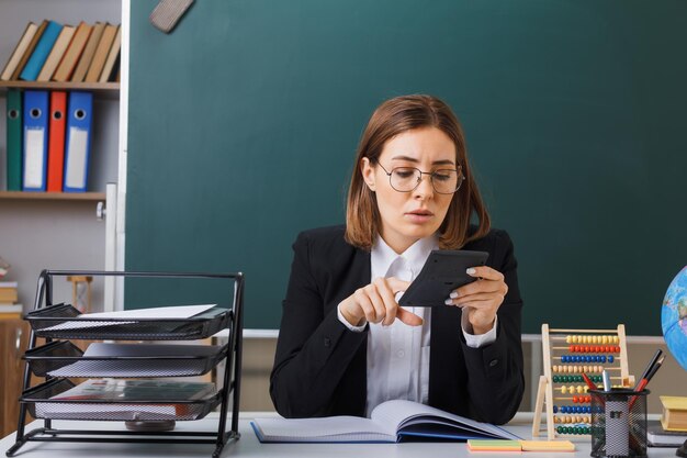 集中して見えるレッスンの準備をしている電卓を使用して教室の黒板の前にある学校の机に座っている眼鏡をかけている若い女性教師