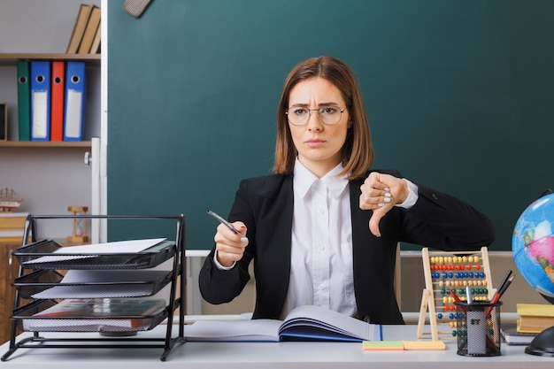 안경을 쓴 젊은 여성 교사는 교실에서 칠판 앞에 앉아 있고, 엄지손가락을 아래로 보여주는 포인터를 들고 수업 기록부를 확인하는 글로브와 함께 칠판 앞에 앉아 있습니다.