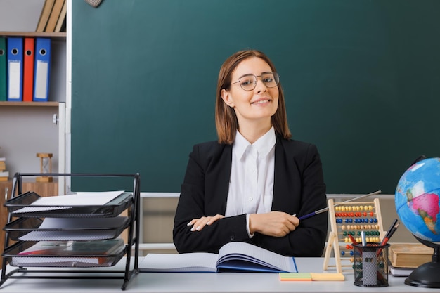 Young woman teacher wearing glasses sitting at school desk in front of blackboard in classroom with abacus and globe checking class register holding pointer smiling confident