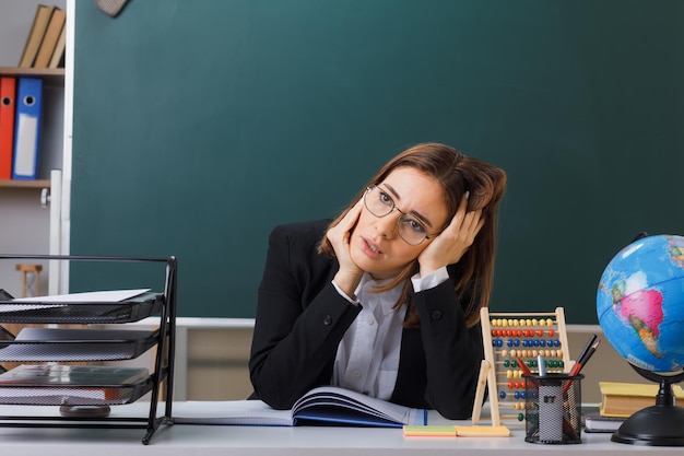 そろばんと地球をチェックするクラスのレジスターが疲れて過労に見える教室の黒板の前にある学校の机に座っている眼鏡をかけている若い女性教師