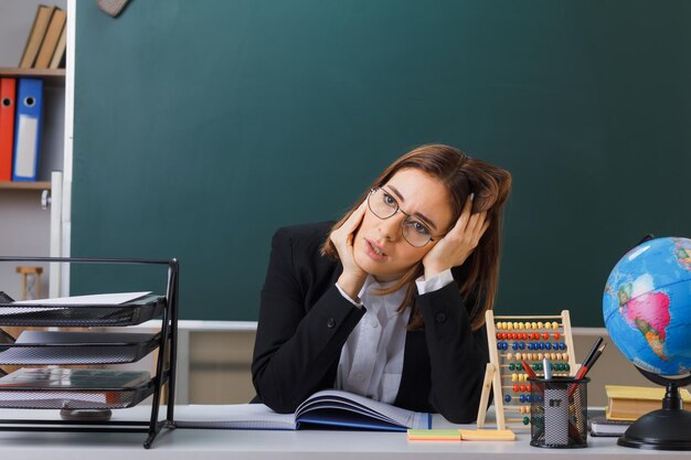 young woman teacher wearing glasses sitting at school desk in front of blackboard in classroom with abacus and globe checking class register looking tired and overworked