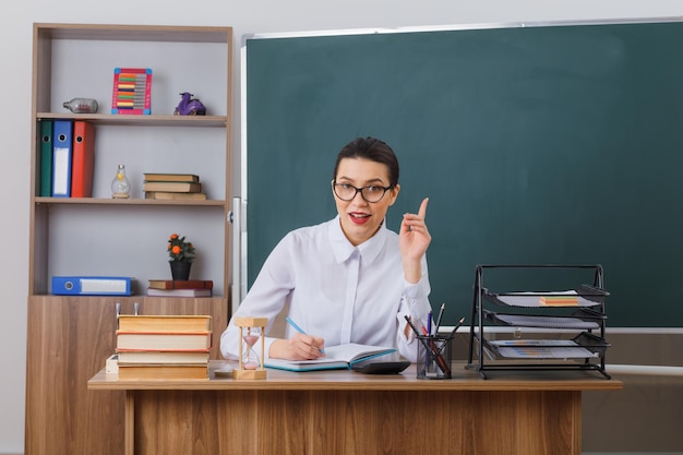 Young woman teacher wearing glasses sitting at school desk in front of blackboard in classroom writing in notebook smiling pointing with index finger up having great idea