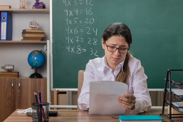 教室の黒板の前で混乱して不機嫌そうに見える空白のページで学校の机に座って眼鏡をかけている若い女性教師