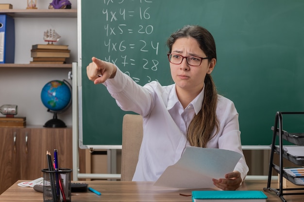 Insegnante di giovane donna con gli occhiali seduto al banco di scuola con pagine vuote che puntano con il dito indice a qualcosa che controlla il lavoro a casa davanti alla lavagna in aula