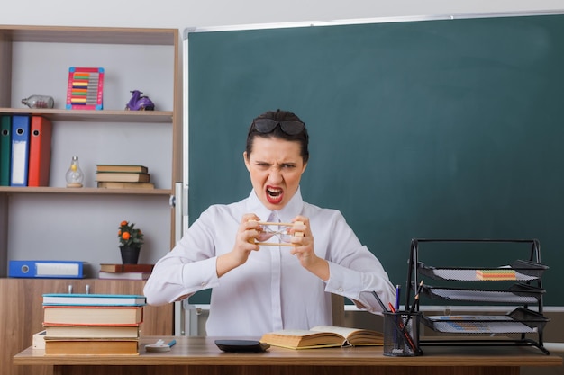Young woman teacher wearing glasses sitting at school desk with book in front of blackboard in classroom holding hourglass angry and frustrated shouting with aggressive expression