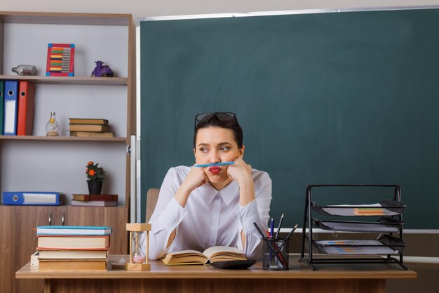 피곤하고 지루해 보이는 입술과 코 사이에 펜을 들고 교실 칠판 앞에 책과 함께 학교 책상에 앉아 안경을 쓴 젊은 여교사