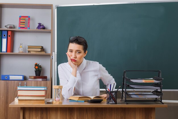 Молодая женщина-учительница в очках сидит за школьной партой с книгой перед доской в классе и смотрит в камеру, уставшая и скучающая, опираясь головой на ладонь