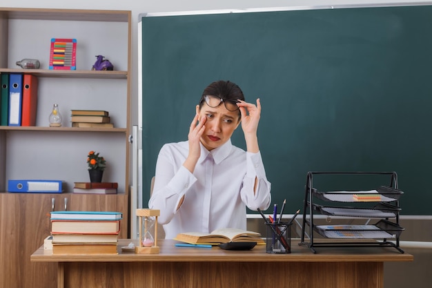 Free photo young woman teacher wearing glasses sitting at school desk with book in front of blackboard in classroom looking tired with sad expression rubbing eye