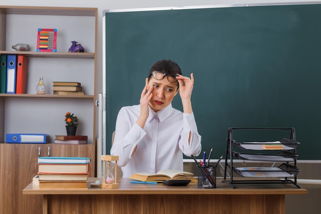 Young woman teacher wearing glasses sitting at school desk with book in front of blackboard in classroom looking tired with sad expression rubbing eye