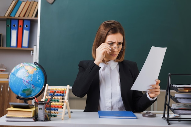 Young woman teacher wearing glasses sitting at school desk with globe and books in front of blackboard in classroom holding white empty sheet of paper looking concentrated