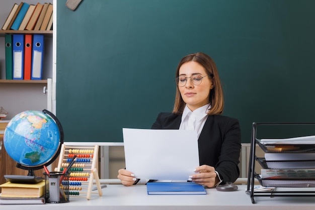 안경을 쓴 젊은 여성 교사는 교실에서 칠판 앞에 글로브와 책을 들고 학교 책상에 앉아 자신감을 보이는 흰색 빈 종이를 들고 있다