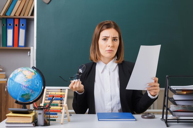 young woman teacher wearing glasses sitting at school desk with globe and books in front of blackboard in classroom holding white empty sheet of paper looking displeased