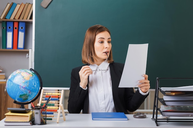 Молодая женщина-учитель в очках сидит за школьной партой с глобусом и книгами перед доской в классе, держа белый пустой лист бумаги, выглядя заинтригованным