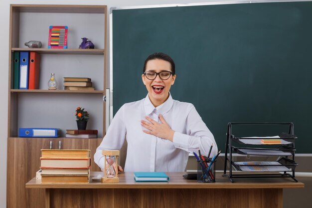 Young woman teacher wearing glasses smiling cheerfully holding hand on her chest feeling thankful sitting at school desk in front of blackboard in classroom