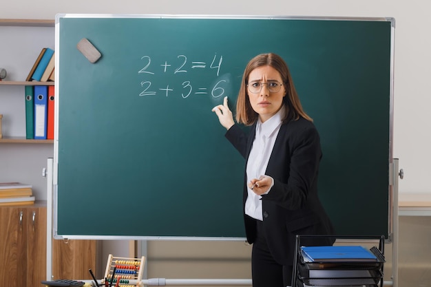 Молодая женщина-учитель в очках, стоящая возле доски в классе, объясняет урок, указывая на доску указателем, выглядя разочарованным
