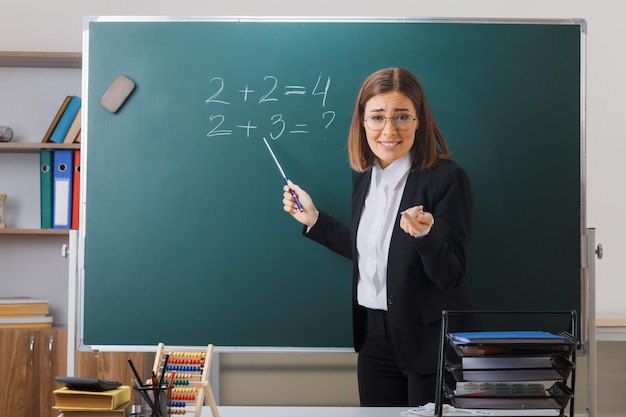 Insegnante di giovane donna con gli occhiali in piedi vicino alla lavagna in classe tenendo il puntatore che spiega la lezione sorridente con espressione scettica