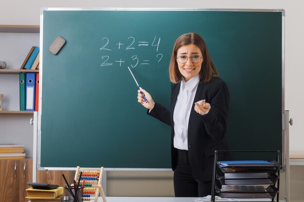 young woman teacher wearing glasses standing near blackboard in classroom holding pointer explaining lesson smiling with skeptic expression