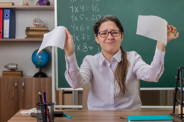 Молодая женщина-учитель в очках разрывает лист бумаги с недовольным видом, сидя за школьной партой перед доской в классе