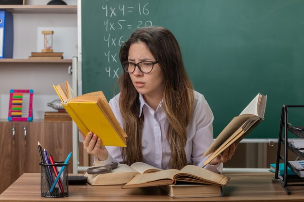 Young woman teacher wearing glasses with books looking confused and very anxious sitting at school desk in front of blackboard in classroom