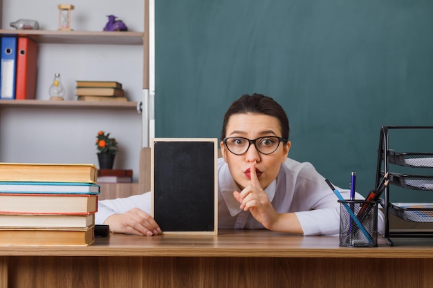 Young woman teacher wearing glasses with small chalkboard sitting at school desk in front of blackboard in classroom making silence gesture with finger on lips