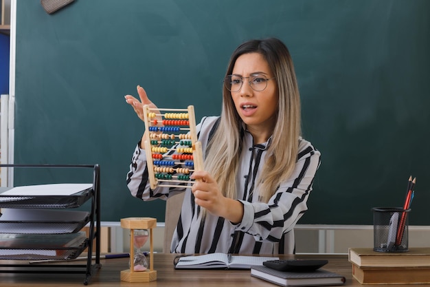 young woman teachersitting at school desk in front of blackboard in classroom explaining lesson holding abacus looking confused and displeased