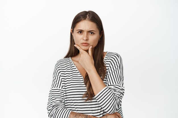 Young woman thinking, having doubts, looking serious at camera with fingers touching chin, ponder decision, white background
