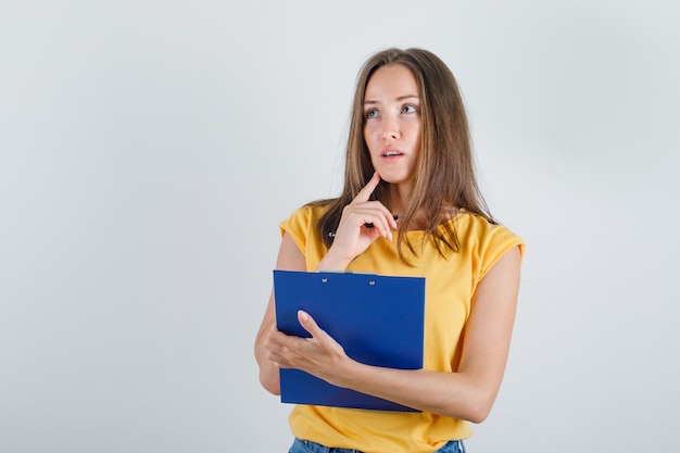 Young woman thinking while holding clipboard in t-shirt, shorts and looking busy