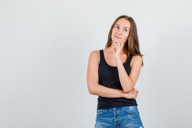 Young woman thinking while looking away in singlet, shorts and looking cheery. front view.