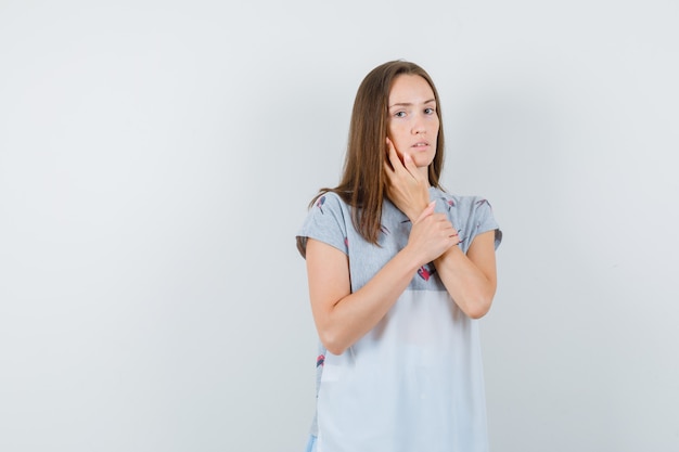 Young woman touching cheek with fingers in t-shirt and looking serious , front view.