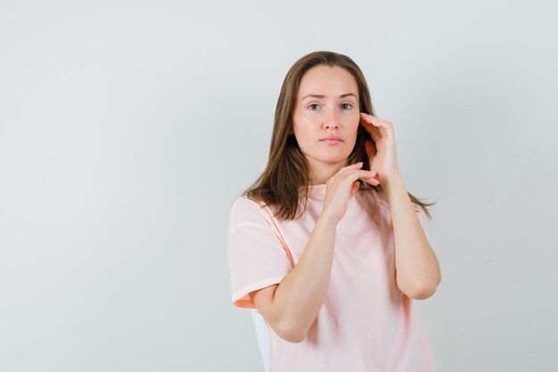 Молодая женщина трогает кожу лица на щеке в розовой футболке и выглядит нежной.