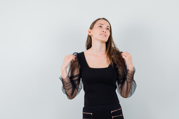 Young woman touching her shoulders while looking away in black blouse and looking pensive. front view.