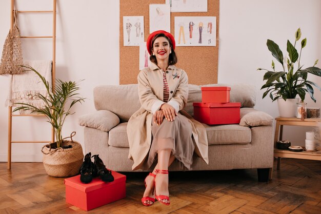 Young woman in trendy autumn outfit poses in cozy room. Pretty woman in fashionable clothes and red heels is sitting on beige sofa next to red boxes.