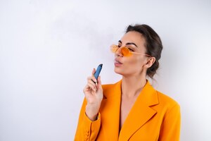A young woman in trendy stylish glasses and a bright orange oversized jacket on a white background smokes an electronic cigarette