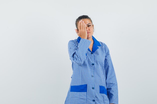 Young woman trying cover eye with hand in blue gingham pajama shirt and looking serious , front view.