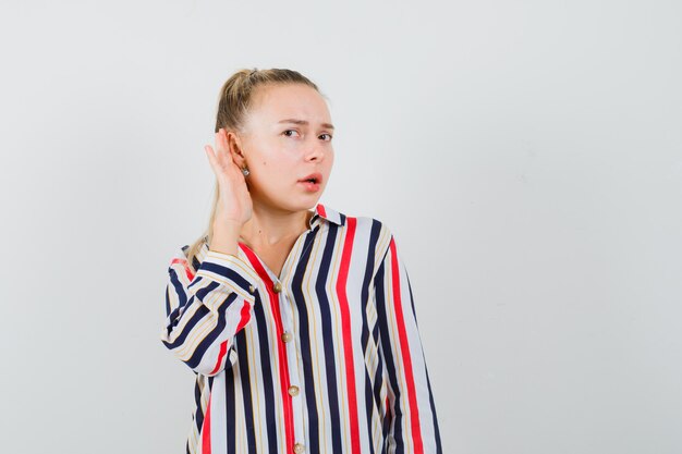Young woman trying to hear something in striped blouse and looking curious and surprised